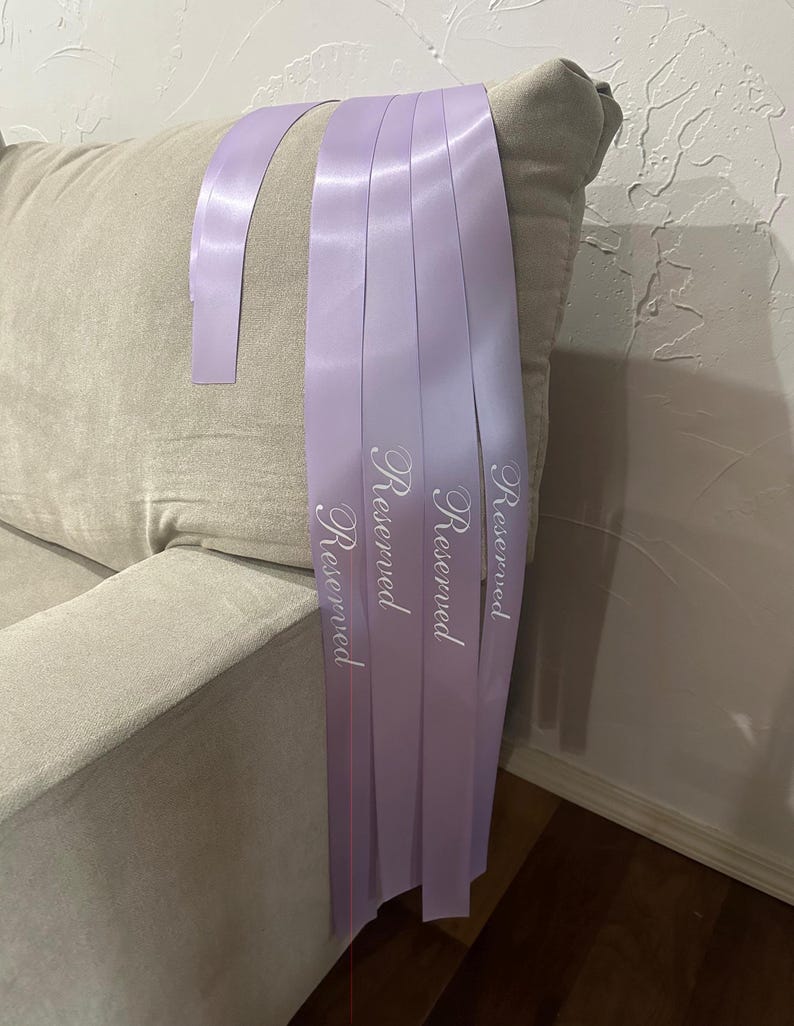 Peut inclure: Plusieurs rubans de satin lavande avec le mot &laquo;&nbsp;Reserved&nbsp;&raquo; imprim&eacute; en &eacute;criture blanche sont drap&eacute;s sur un canap&eacute; beige. Les rubans sont dispos&eacute;s en cascade, avec les extr&eacute;mit&eacute;s des rubans visibles.