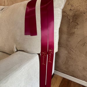 Peut inclure: Rubans de satin bordeaux avec le mot "Reserved" en script dor&eacute;. Les rubans sont drap&eacute;s sur le bras d'un canap&eacute; beige. L'arri&egrave;re-plan est un mur brun textur&eacute;.
