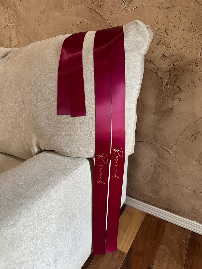 Peut inclure: Rubans de satin bordeaux avec le mot "Reserved" en script dor&eacute;. Les rubans sont drap&eacute;s sur le bras d'un canap&eacute; beige. L'arri&egrave;re-plan est un mur brun textur&eacute;.
