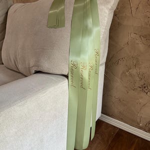 Peut inclure: Ruban de satin vert clair avec le mot "Reserved" imprim&eacute; en lettres dor&eacute;es, drap&eacute; sur le bras d'un canap&eacute; beige. Plusieurs rubans sont visibles, avec une petite pile de rubans sur le coussin du canap&eacute;.