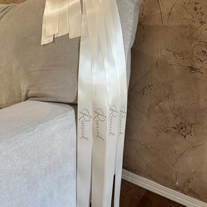 Peut inclure: Rubans en satin ivoire avec le mot "Reserved" imprim&eacute; dans une &eacute;criture d&eacute;licate. Les rubans sont drap&eacute;s sur une chaise rembourr&eacute;e grise, sugg&eacute;rant un th&egrave;me de mariage ou d'&eacute;v&eacute;nement. Les rubans mesurent environ 5 cm de large.