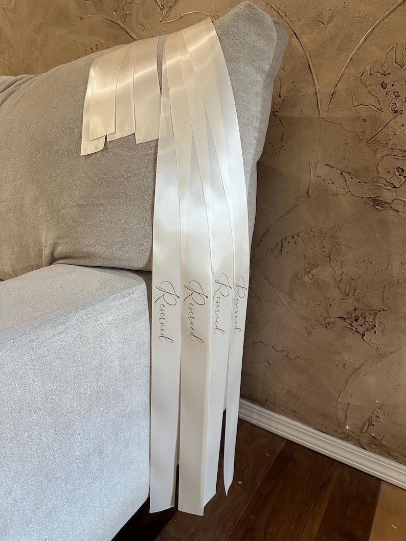 Peut inclure: Rubans en satin ivoire avec le mot "Reserved" imprim&eacute; dans une &eacute;criture d&eacute;licate. Les rubans sont drap&eacute;s sur une chaise rembourr&eacute;e grise, sugg&eacute;rant un th&egrave;me de mariage ou d'&eacute;v&eacute;nement. Les rubans mesurent environ 5 cm de large.