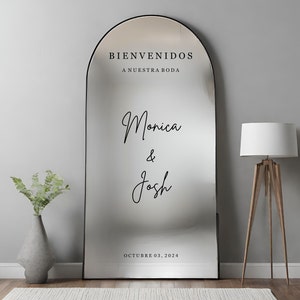 May include: A black arched mirror with a welcome sign in Spanish that reads "Bienvenidos a nuestra boda" and the names "Monica & Josh" written in cursive. The date "Octubre 03, 2024" is written below the names.