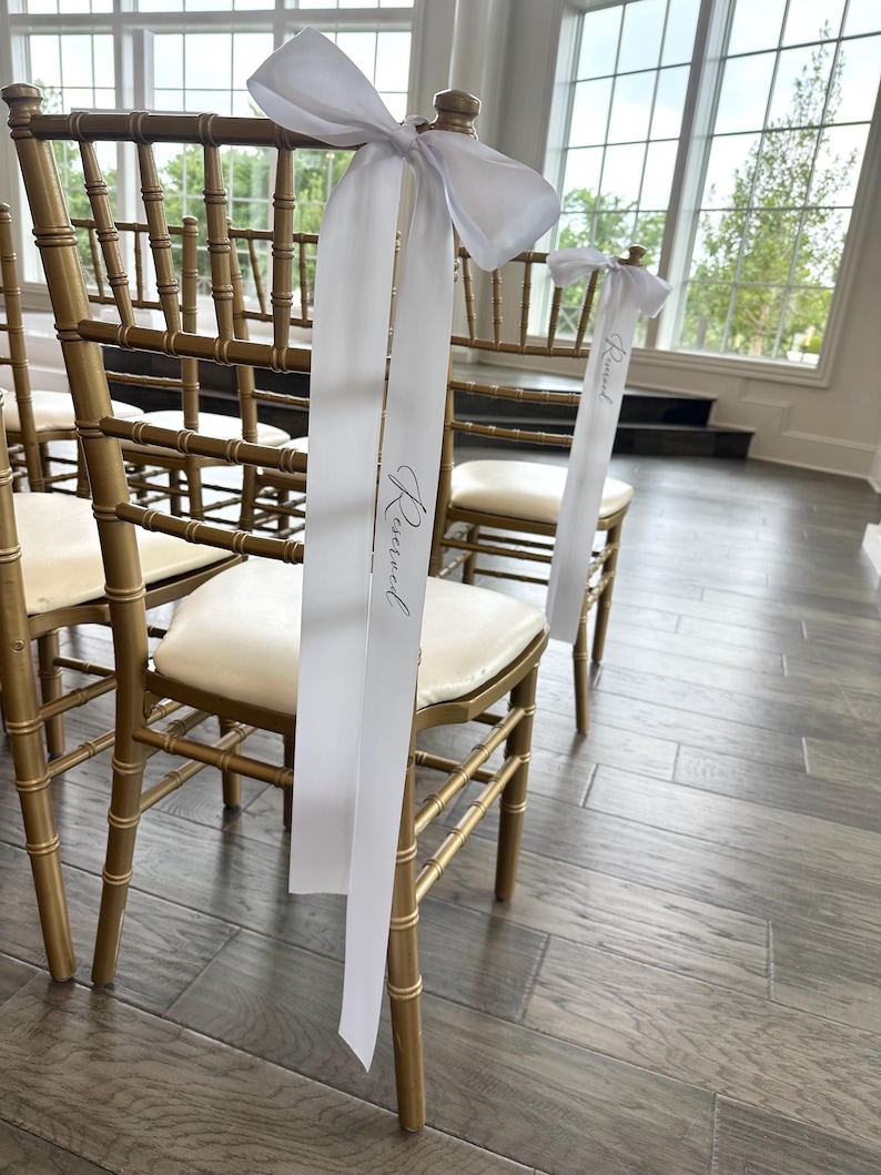 Peut inclure: Rang&eacute;e de chaises en bois dor&eacute; avec des assises cr&egrave;me. Des rubans blancs, nou&eacute;s en n&oelig;uds, sont attach&eacute;s aux dossiers des chaises, avec le mot "Reserved" imprim&eacute;. Les chaises sont dans une pi&egrave;ce avec de grandes fen&ecirc;tres.