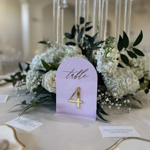 May include: An arched light pink acrylic table number sign featuring the word Table in black script and a large gold number 4. The sign stands upright on a base, positioned in front of a floral centerpiece with white flowers and dark green leaves.