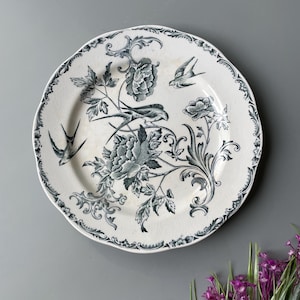 Könnte beinhalten: Weißer Keramikteller mit gewelltem Rand, verziert mit einem floralen und Vogeldesign in Grau- und Weißtönen. Der Teller hat einen detaillierten Rand und eine Vintage-Ästhetik, geeignet zum Servieren von Speisen oder als dekoratives Stück.
