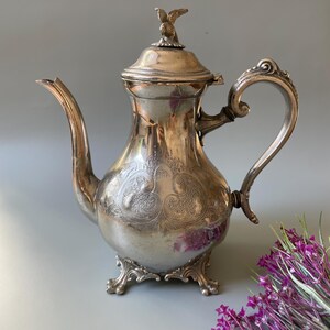 May include: An antique silver-plated coffee pot with ornate detailing. The pot features a bird-shaped finial on the lid, a curved spout, and an elaborately designed handle. The body of the pot is decorated with floral patterns and rests on decorative feet.