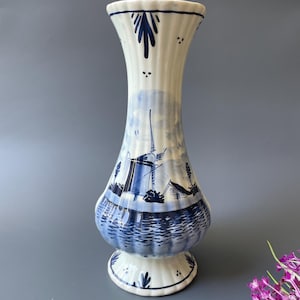 May include: A decorative ceramic vase in white and blue, featuring a traditional Dutch windmill design. The vase has a flared top and a rounded body, standing on a pedestal base. The blue design depicts a windmill and water scene.