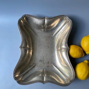 Pode incluir: Uma bandeja de servir vintage em tons de prata com uma borda decorativa e ondulada. A bandeja tem uma forma retangular com laterais curvas e um acabamento ligeiramente desgastado. Três limões amarelos brilhantes estão dispostos sobre um fundo azul.