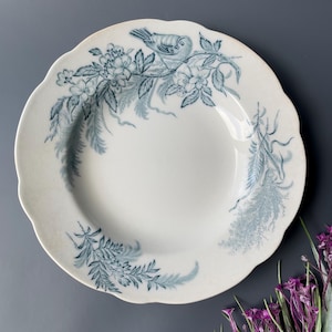 Puede incluir: Plato de cerámica blanca con borde festoneado, adornado con un diseño floral y de pájaros azules. El plato presenta un pájaro central posado en una rama rodeado de flores y follaje. Una pieza decorativa clásica.