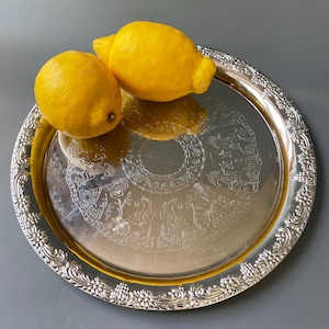 Peut inclure: Deux citrons jaunes sur un plateau de service en argent avec un motif orné. Le plateau a un bord surélevé et un motif floral.