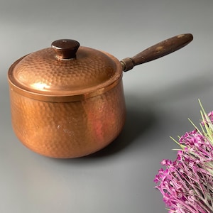 May include: A copper saucepan with a hammered finish, a wooden handle, and a matching lid. The pot is a warm copper color and appears to be vintage. The handle and lid knob are made of dark wood. The pot is designed for cooking.