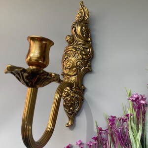 May include: Ornate gold-colored wall sconce with a curved arm and a candle holder. The sconce features detailed floral and scrollwork designs. The background is a light gray wall with purple flowers.