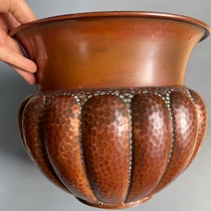 May include: A copper-coloured, hammered metal planter with a wide, flared rim. The lower portion features a series of vertical, rounded panels with a textured, dimpled surface. The planter has a decorative, antique appearance.