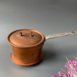 May include: A copper saucepan with a lid and a long, silver-toned handle. The pot sits on a matching copper base. The copper has a warm, reddish-brown hue, and the handle shows signs of wear.
