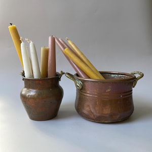 Peut inclure: Deux pots en cuivre avec des poignées en laiton, l'un haut et étroit et l'autre large et peu profond, contenant chacun une collection de bougies effilées dans des tons jaune, blanc et rose poudré avec des mèches visibles au sommet.
