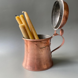 Puede incluir: Una jarra de cobre con tapa abatible y asa curva, que presenta un interior metálico. Tres velas cónicas de cera de abeja de color amarillo claro están colocadas dentro del recipiente, sobresaliendo por la parte superior abierta.