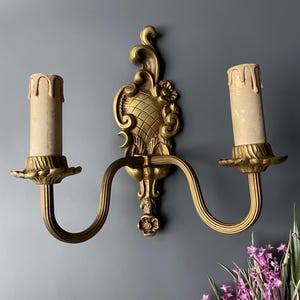 May include: A gold-toned wall sconce with two candle-style light bulbs. The sconce has a decorative, ornate design with a floral motif.