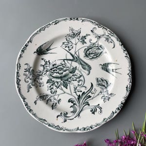 Puede incluir: Plato de cerámica blanca con borde festoneado, con un diseño floral y de pájaros en tonos verdes. El plato tiene un borde detallado y un motivo floral central. El diseño incluye flores y pájaros estilizados.