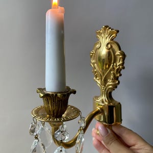 May include: A gold-toned wall sconce featuring an ornate backplate with scrollwork details, a curved arm, and a candle cup. A white taper candle with a lit flame sits in the cup, while clear, faceted crystal teardrop prisms hang below.