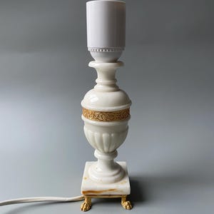 Może przedstawiać: Kremowa lampa stołowa z białym abażurem. Lampa ma dekoracyjny korpus w kształcie urny ze złotą opaską i stoi na kwadratowej podstawie ze złotymi nóżkami w kształcie pazurów. Biały przewód wychodzi z podstawy.