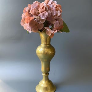 Op de afbeelding: Een hoge, antieke messing vaas houdt een boeket perzik hydrangeas vast. De vaas heeft een uitlopende basis, een bolvormig middengedeelte met geëtste details en een gegolfde bovenkant. De bloemen zijn een mix van lichtroze en perziktinten, met groene bladeren.