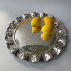 Peut inclure: Un plateau de service rond de couleur argentée avec un bord festonné décoratif présentant des motifs découpés complexes. Deux citrons jaune vif sont posés sur la surface réfléchissante du plateau, créant un reflet clair du fruit.