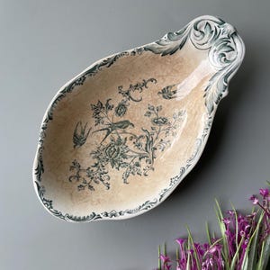Può includere: Un piatto ovale in ceramica con finitura smaltata beige craquelé, caratterizzato da motivi a trasferimento verde scuro raffiguranti uccelli, fiori e fogliame. Il bordo è decorato con motivi botanici ornamentali e bordi smerlati.