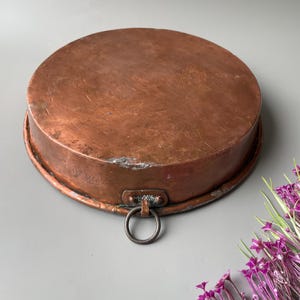 Puede incluir: Una sartén redonda de cobre antiguo con una pátina oscura. Tiene un borde elevado y un asa con un anillo. El cobre presenta zonas de desgaste y decoloración. Flores moradas en la esquina inferior derecha.