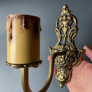 Könnte beinhalten: Eine goldfarbene Wandleuchte mit einem verzierten Design. Die Leuchte hat einen einzelnen Kerzenhalter und einen gebogenen Arm. Der Kerzenhalter ist leer.