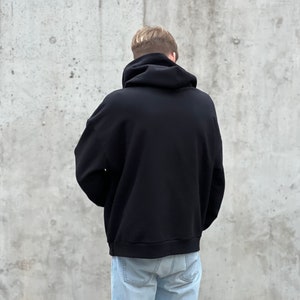 K&ouml;nnte beinhalten: Ein schwarzer Kapuzenpullover mit einer Kordelzugkapuze. Der Pullover wird von einer Person getragen, die vor einer Betonwand steht.