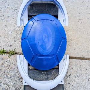 May include: A blue, geometric-patterned disc sits atop a gray and black electric skateboard. The skateboard has a white frame and a black footpad. The disc appears to be a protective cover or accessory for the skateboard.