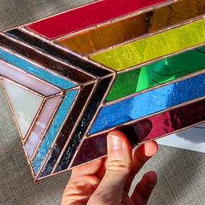 Puede incluir: Una bandera del orgullo de vidrieras con rayas rojas, naranjas, amarillas, verdes, azules y moradas. La bandera se sostiene cerca de un papel con contornos de bandera y una pegatina de descarga digital.