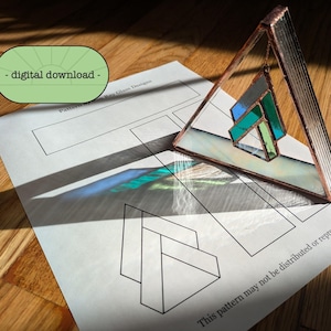 May include: A stained glass triangle with a geometric pattern in shades of blue and green. The triangle is hanging from a copper wire and is casting a shadow on a white sheet of paper with a pattern printed on it. The paper has the text "This pattern may not be distributed or reproduced" printed on it.