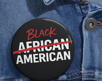 May include: A round pinback button with a black background. The text reads 'BLACK' in red script, followed by 'AFRICAN' and 'AMERICAN' in white block letters. A red line strikes through the word 'AFRICAN'. The button is attached to blue denim fabric.