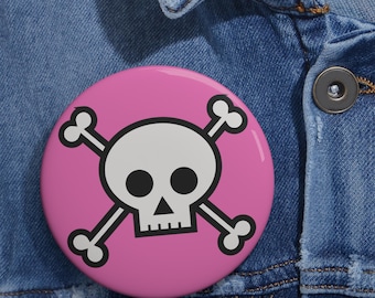 May include: A circular pin-back button featuring a graphic of a white skull and crossbones with black outlines set against a solid pink background. The design is centered on the button, which has a smooth, glossy finish and a rounded edge.