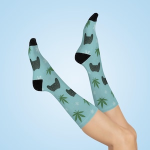 May include: Light blue socks with black heels and toes, featuring a pattern of black video game controllers, green cannabis leaves, and white snowflakes. The socks are displayed against a light blue background.