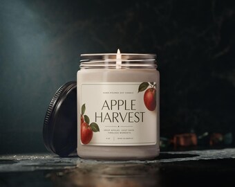 May include: A clear glass jar candle with a lit wick and a black lid placed to the side. The label features the text APPLE HARVEST, illustrations of red apples with green leaves, and the text 8 OZ, MADE IN AMERICA, CRISP APPLES, COZY DAYS, TIMELESS MOMENTS.