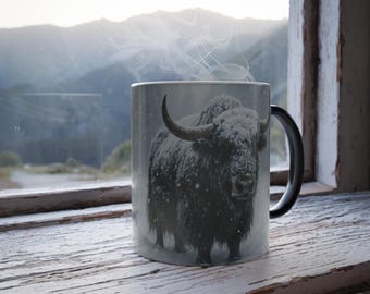 May include: A white ceramic mug featuring a black and white image of a yak covered in snow. Steam rises from the mug, suggesting a hot beverage. The mug is set against a backdrop of a window with a mountain view.