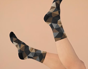 May include: A pair of crew-length socks featuring a geometric pattern composed of triangular shapes in shades of black, dark gray, light gray, and tan. The socks have solid black cuffs, heels, and toe sections, providing a consistent look.