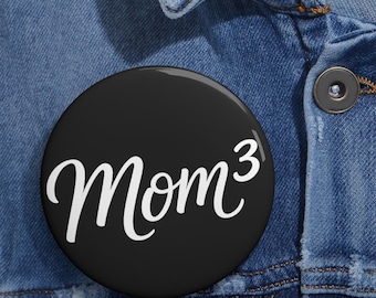 May include: A circular black pin-back button featuring the white text "Mom³" in a cursive script font. The button is attached to a blue denim fabric background with a visible metal button and stitching details.