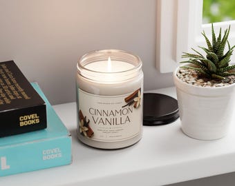 May include: A clear glass jar candle with a white label reading Cinnamon Vanilla, featuring a lit wick and a black lid. The label displays illustrations of cinnamon sticks and vanilla flowers. The candle is positioned next to two books and a small succulent.