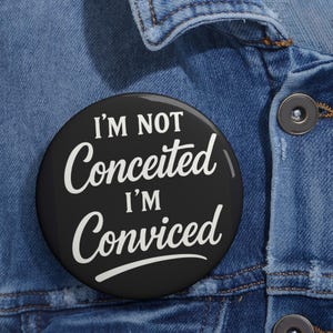 May include: A round black pin-back button featuring white text that reads I'M NOT Conceited I'M Conviced in a mix of bold sans-serif and cursive script fonts. The button is attached to a blue denim fabric surface with visible metal buttons.