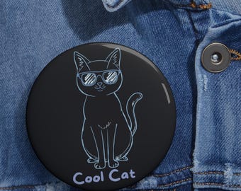 May include: A round black pin-back button featuring a light blue line art illustration of a cat wearing sunglasses. The cat is centered on the button, and the text "Cool Cat" is printed in light blue letters at the bottom of the circular design.