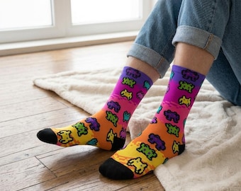 May include: Colorful socks with a pixelated video game character pattern. The socks feature a gradient of colors, including purple, pink, orange, and yellow, with black toes and heels. The socks are worn with blue jeans.