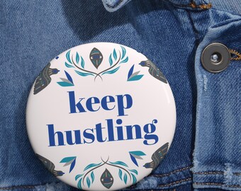 May include: A round pinback button with a white background featuring the blue text keep hustling in the center. The design includes symmetrical teal and dark blue leaf patterns and stylized bird illustrations positioned around the text.