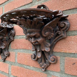 May include: Two ornate, cast iron wall sconces with a dark brown finish are mounted on a red brick wall. Each sconce features a detailed face with wings and a circular shelf, perfect for displaying small items.