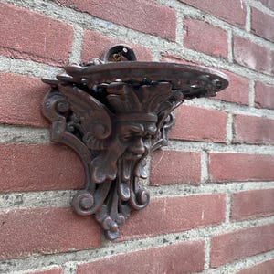 May include: Ornate, dark brown cast iron wall sconce with a decorative face and wings. The sconce has a small, round shelf and is mounted on a red brick wall. The design is detailed and classical.