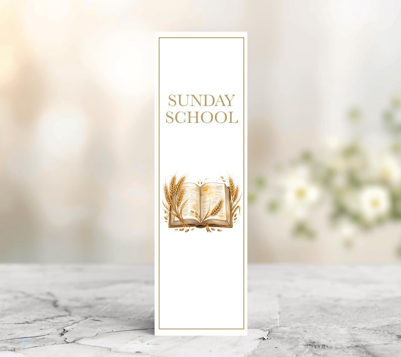 May include: A white bookmark featuring the text "SUNDAY SCHOOL" in gold. An illustration of an open book with wheat surrounds the text. The bookmark has a gold border and is on a marble surface.
