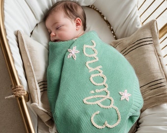 May include: A baby sleeping in a wicker bassinet, swaddled in a green knitted blanket with the word "Eliana" embroidered in white. The blanket has pink flower accents.
